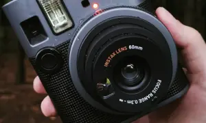 Szary aparat natychmiastowy Instax z obiektywem 60mm, trzymany w dłoni na tle lasu.