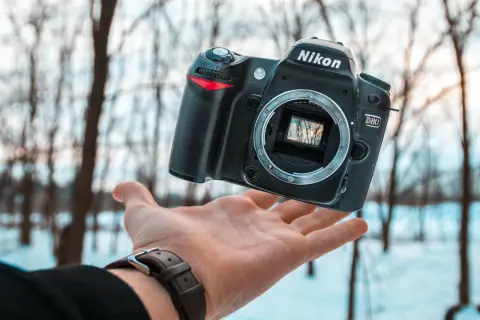 Oto jak wygląda aparat fotograficzny Nikon D80, unoszący się nad dłonią na tle zimowego lasu.