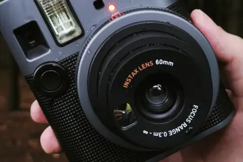 Szary aparat natychmiastowy Instax z obiektywem 60mm, trzymany w dłoni na tle lasu.