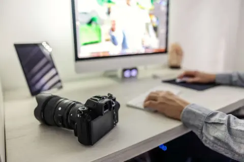 Aparat fotograficzny leży obok tabletu, a osoba przy komputerze pracuje nad zdjęciami. To pokazuje, jak zgrać zdjęcia z aparatu na laptop.