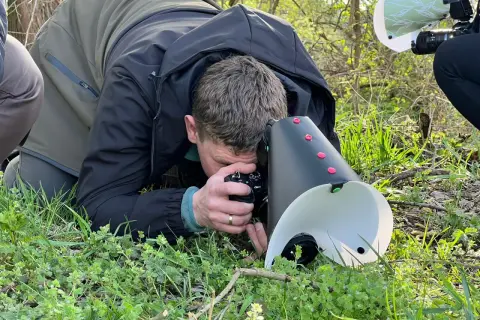 Fotograf z aparatem i dyfuzorem do makro pochyla się nad trawą, by uchwycić drobne detale natury.