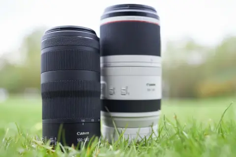Dwa obiektywy Canon 100-400, jeden czarny, drugi biały, stoją w trawie.